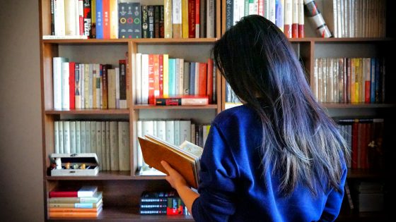 A person with long hair is standing in front of a full bookshelf, holding and reading an open book