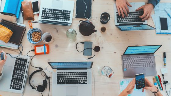 Top-down view of a collaborative tech workspace with multiple laptops, smartphones, notebooks, and people working together around a large desk
