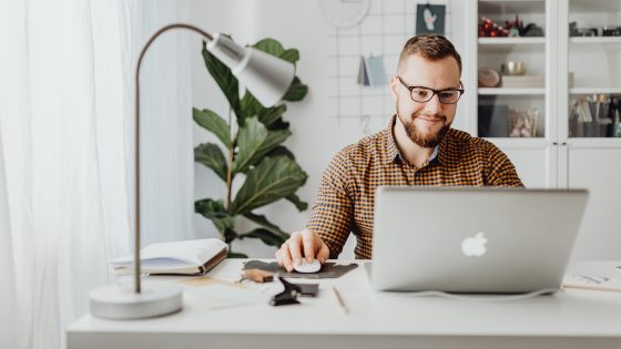 Smiling man working from home at a modern desk with a laptop, using a wireless mouse in a bright, minimalist home office with indoor plants and bookshelves in the background
