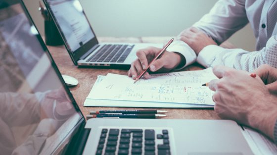 Two professionals analyzing documents and taking notes with laptops on a desk during a business planning meeting