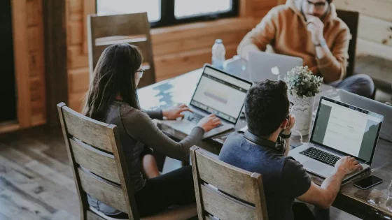 Three people are working on their laptops at a wooden table in a well-lit room, possibly collaborating on a project