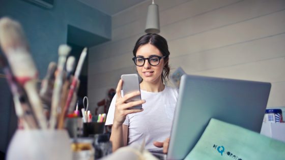 A woman wearing glasses is sitting at a desk with a laptop, looking at her phone, while surrounded by various office and art supplies