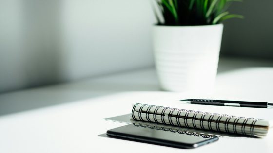 A phone and a spiral notebook with a pen are resting on a clean, sunlit desk next to a white potted plant