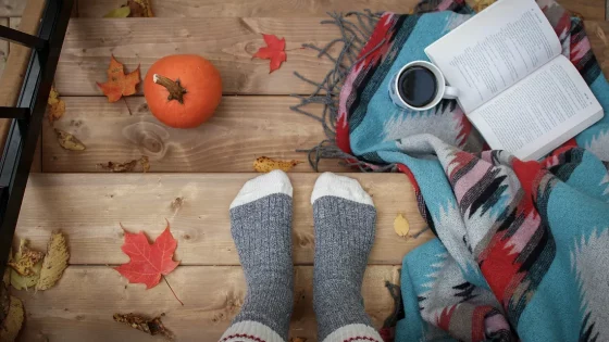 A person's feet in cozy socks are shown on a wooden deck with autumn leaves, a small pumpkin, a patterned blanket, a cup of coffee, and an open book