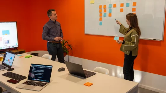 A woman is pointing at sticky notes on a whiteboard, while a man stands nearby, both participating in a meeting in a bright orange room