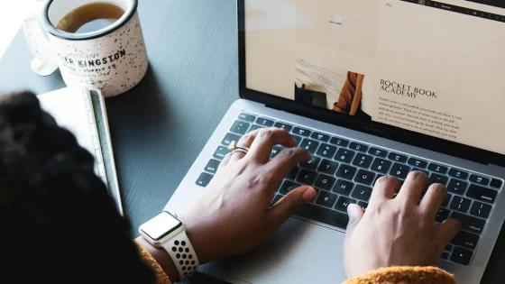 A person is typing on a MacBook, with a cup of coffee and a notebook on a dark desk