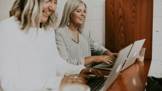 Two women are smiling and working on their laptops at a wooden counter, with one in the foreground slightly out of focus and the other looking at her screen