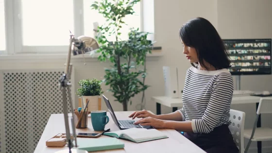 A woman is sitting at a well-lit desk, typing on a laptop with a mug and notebook nearby, with a second monitor displaying images in the background