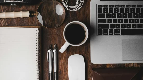 A wooden desk with a laptop, a magnifying glass, a notebook, pens, a cup of coffee, and other miscellaneous work items