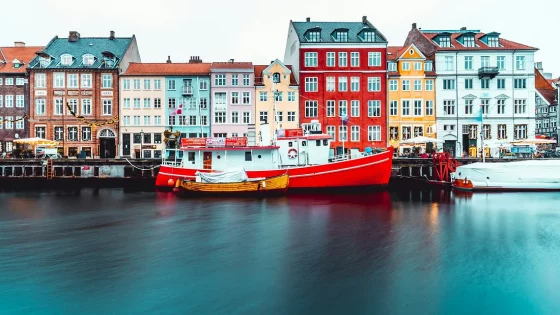 A long-exposure shot of a harbor in Copenhagen, showcasing a row of colorful buildings and two boats moored in the water