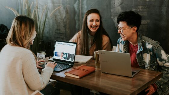Three freelancers sitting at a wooden table, smiling and working together on laptops, discussing project plans and collaborating on ideas in a casual office setting