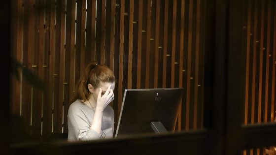 A woman with red hair, tied back in a ponytail, sits at a desk in a dimly lit room. Her head is bowed and her right hand is covering her face, suggesting she is experiencing stress or exhaustion. In front of her is a computer monitor with a lit-up screen. The background features a textured wall made of wooden slats. The image is seen from a distance through a window or a similar partition.