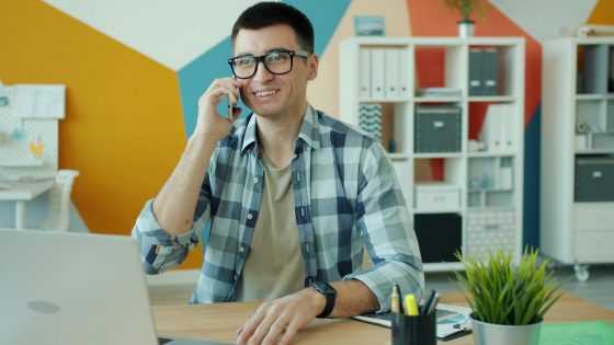 Smiling man wearing glasses and a checkered shirt talking on the phone while sitting at a desk with a laptop, notebook, and plant in a brightly colored modern office