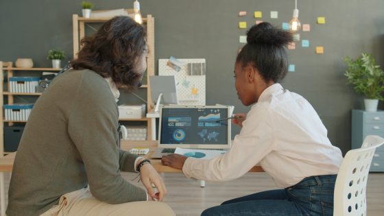 Alt text: Two coworkers reviewing data charts on a laptop at a desk in a modern office