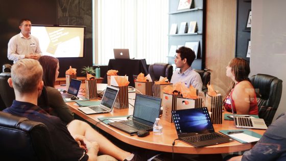 A group of professionals sitting around a large oval conference table during a presentation. A man stands at the front of the room next to a display screen showing a presentation. The team members listen attentively with open laptops, notebooks, and gift bags arranged in front of them on the table.