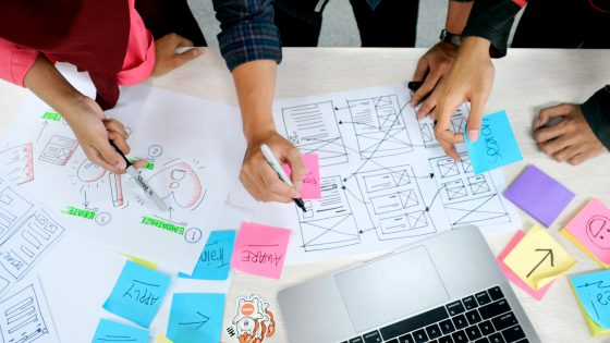Team members collaborating around a table covered in wireframe sketches, sticky notes, and a laptop during a digital campaign planning session